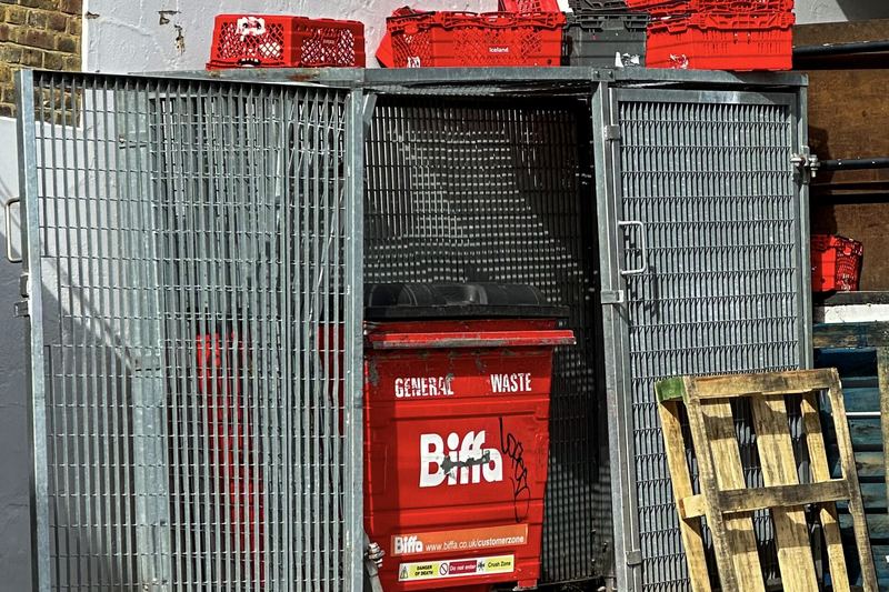 Biffa general waste skip with red plastic storage crates stacked on metal cage units and wooden pallet at Iceland Motor Lane service yard, Gillingham, Kent, Eng